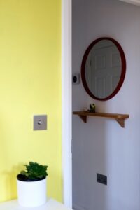 Yellow accent wall with round mirror and wooden shelf in intentionally designed home - Rhizo Home Feng Shui